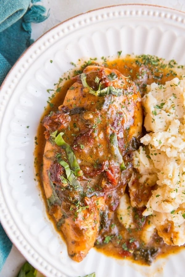 Baked Creamy Sun-Dried Tomato Chicken on a white plate with a side of mashed potatoes. A blue napkin to the side and fresh basil.