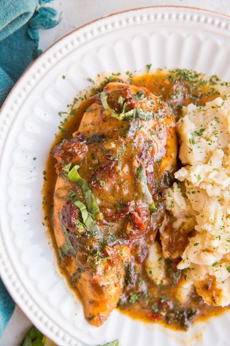Baked Creamy Sun-Dried Tomato Chicken on a white plate with a side of mashed potatoes. A blue napkin to the side and fresh basil.