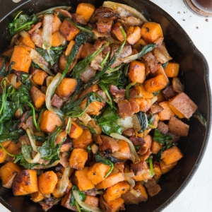 Sweet Potato Hash Recipe in a skillet with crispy bacon, onions, and spinach and a linen wrapped around the handle of the skillet. Pepper grinder to the side.