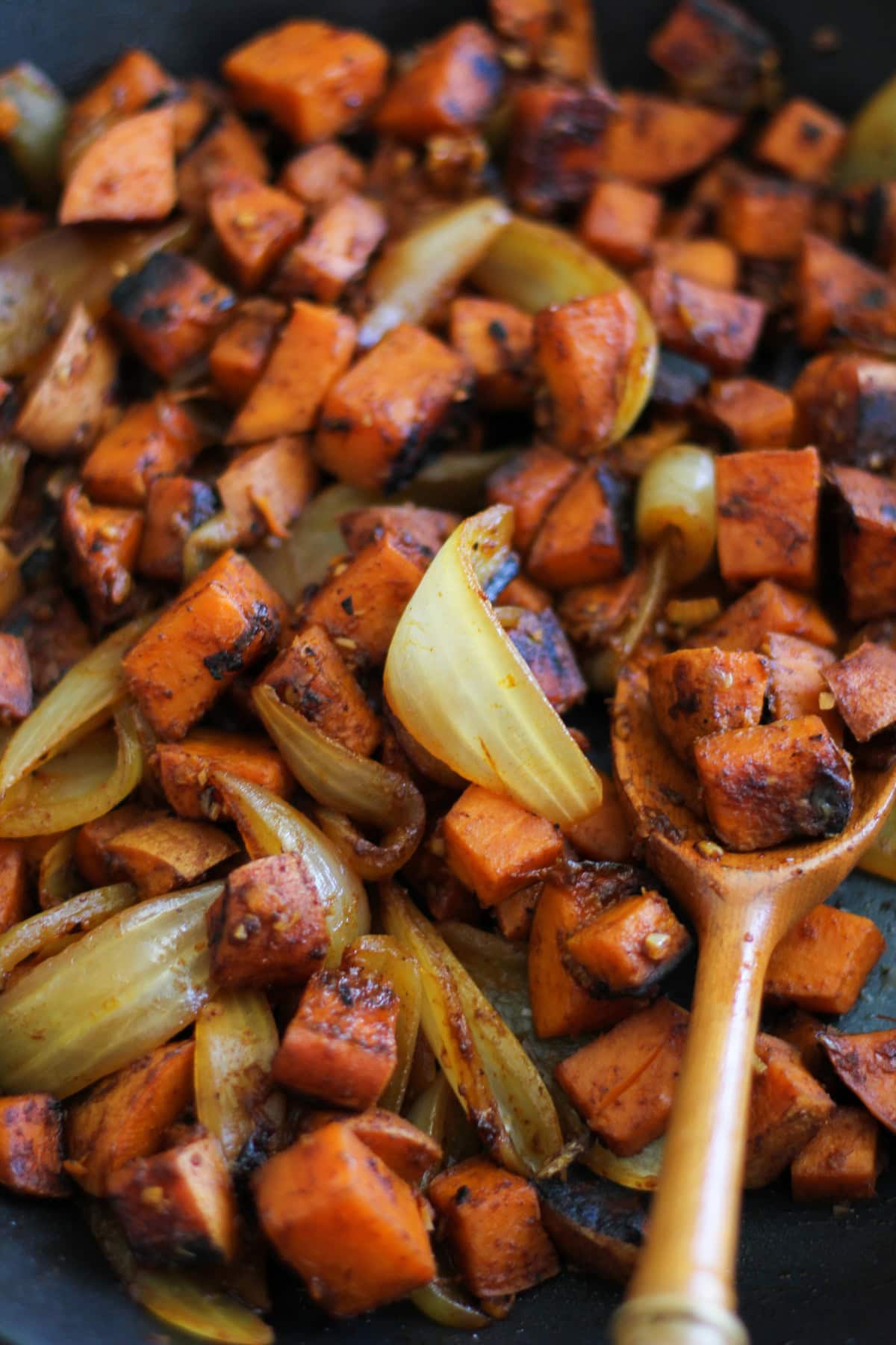 A close-up of sweet potato home fries and caramelized onion wedges being stirred with a wooden spoon in a pan. The well-seasoned vegetables are slightly charred, highlighting the delicious blend of flavors.