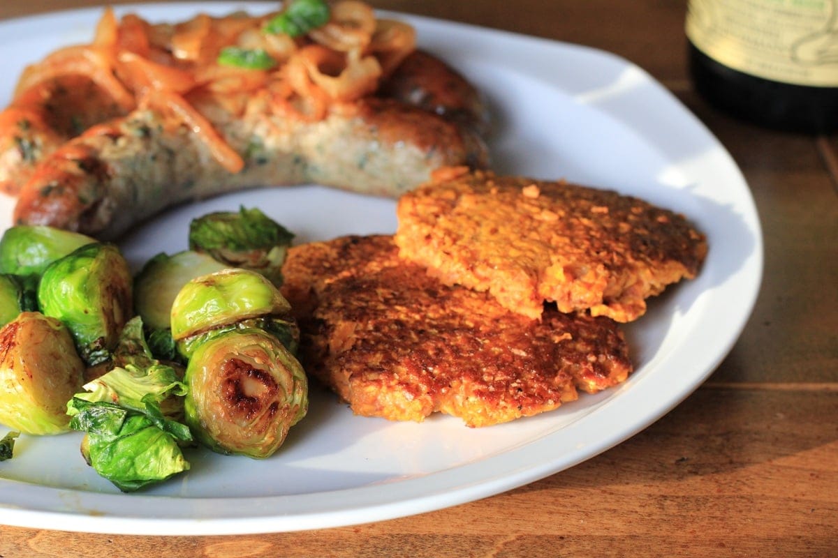 Sweet Potato hash browns on a plate with two sausages topped with caramelized onions and a side of brussel sprouts.