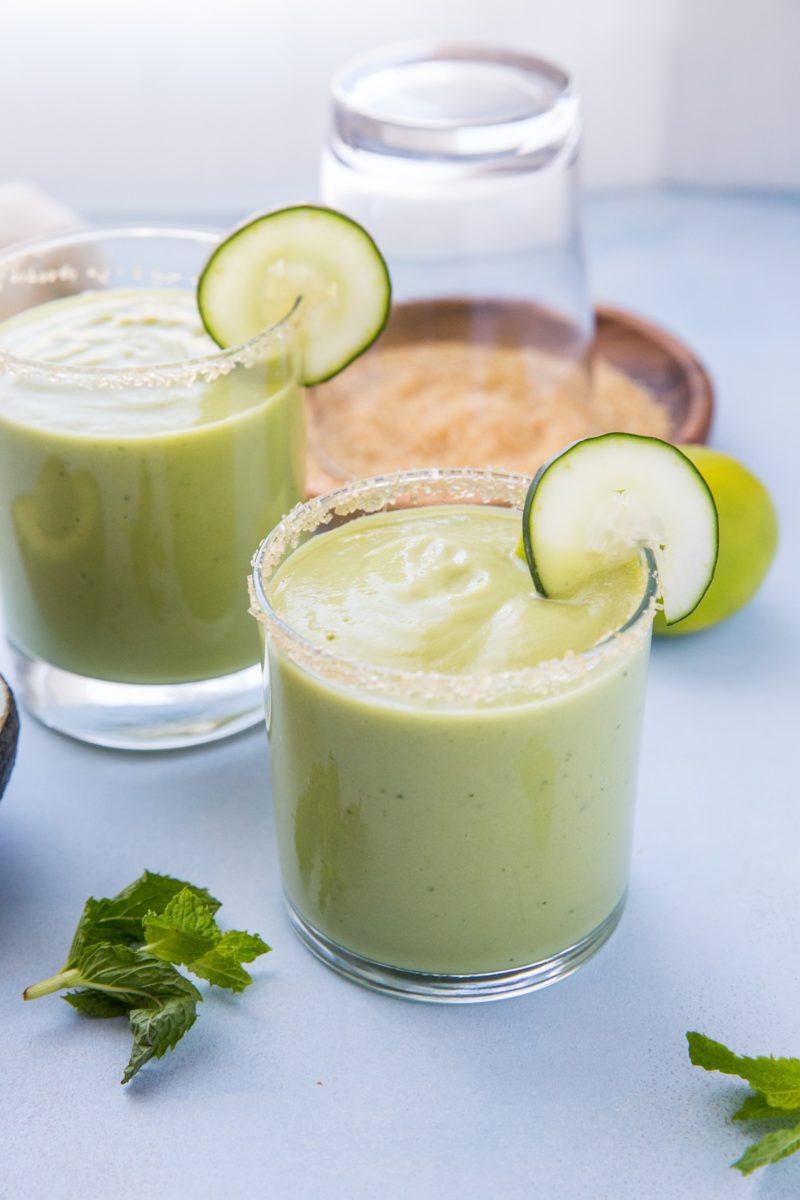 Two glasses of avocado cocktail on a blue surface with fresh mint leaves all around.