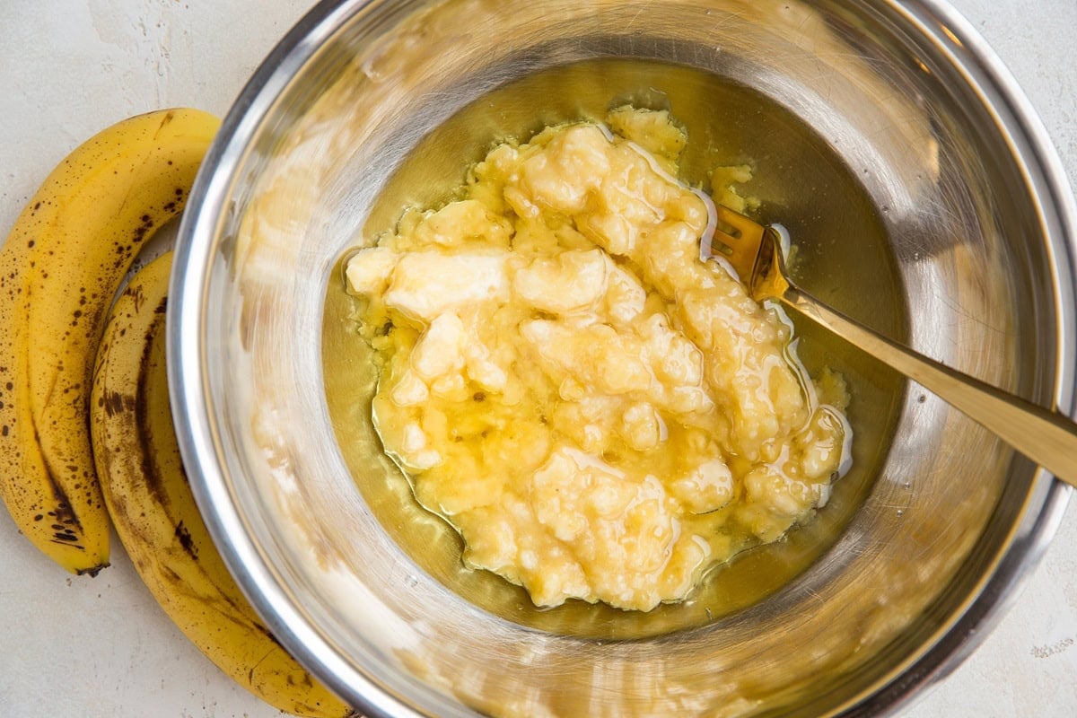 A metal bowl with mashed bananas and a fork inside, next to two ripe bananas on a light-colored surface—perfect for starting your Vegan Banana Bread recipe.