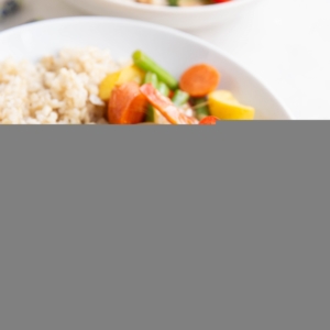 Big white bowl of vegetable stir fry with peanut sauce and brown rice with an orange napkin and blue chopsticks to the side.