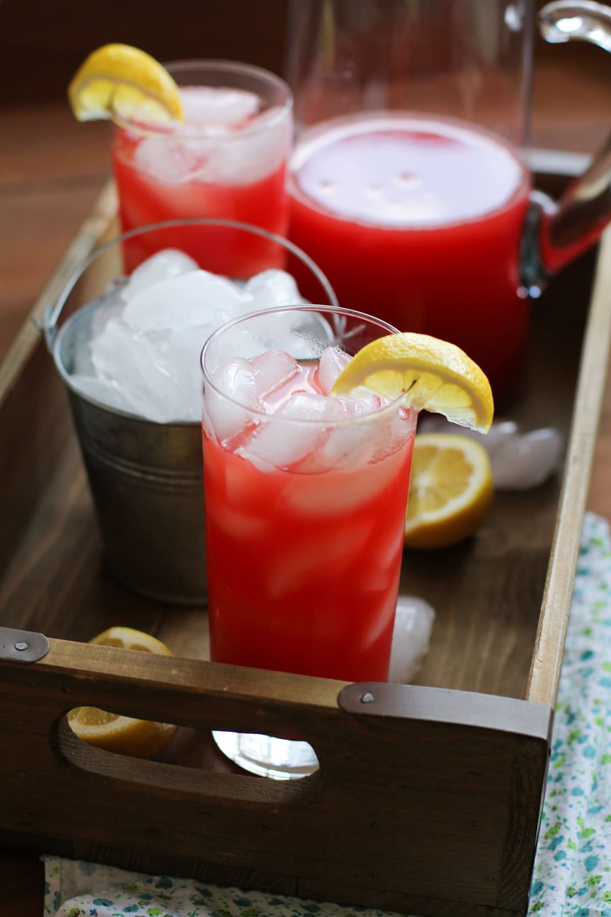 Wooden tray with two glasses and a pitcher of pink Watermelon Agua Fresca with ice and a floral napkin. Ready to serve.