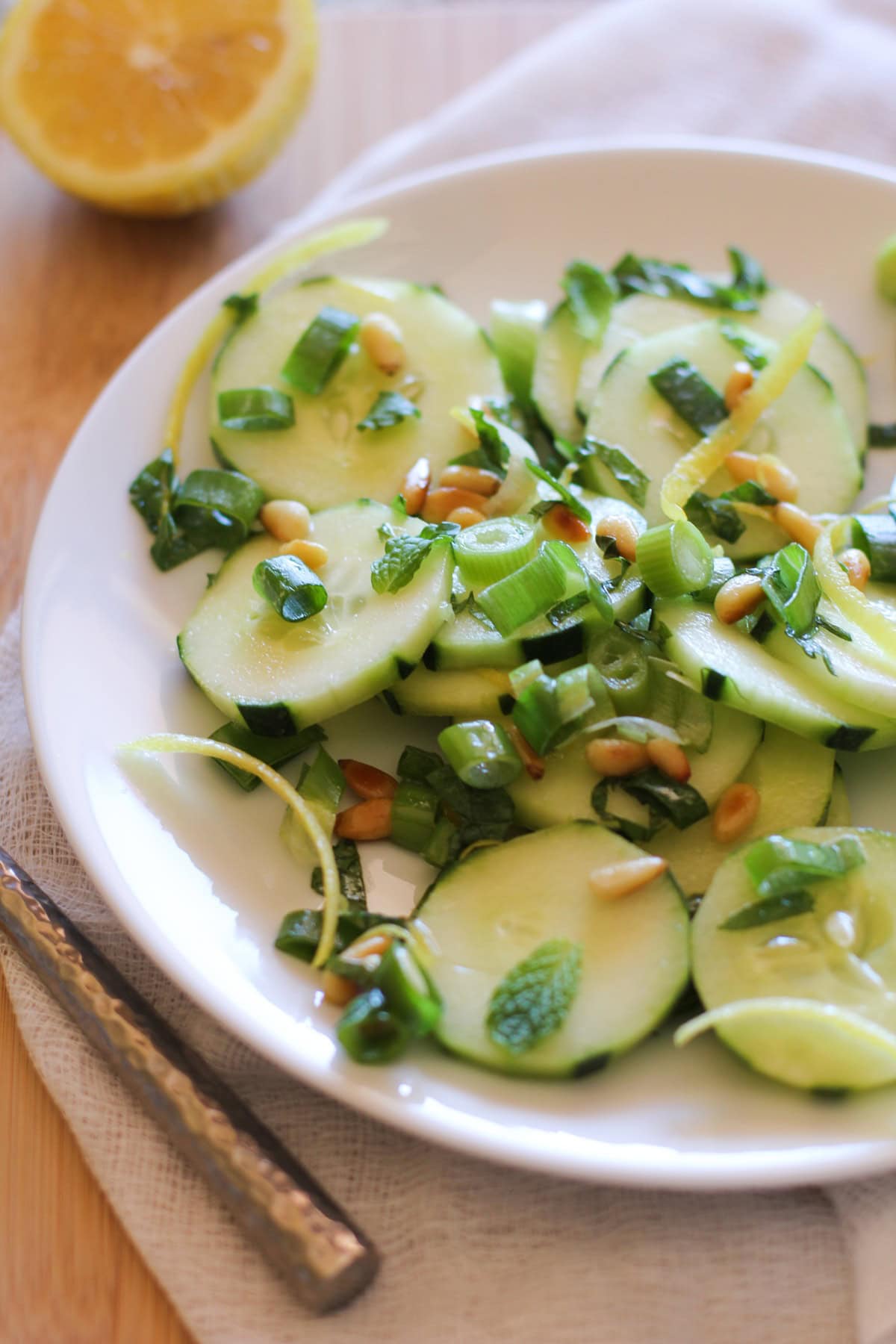 Sliced cucumbers on a plate with lemon zest, mint, green onions, and pine nuts with a fork to the side, ready to serve.
