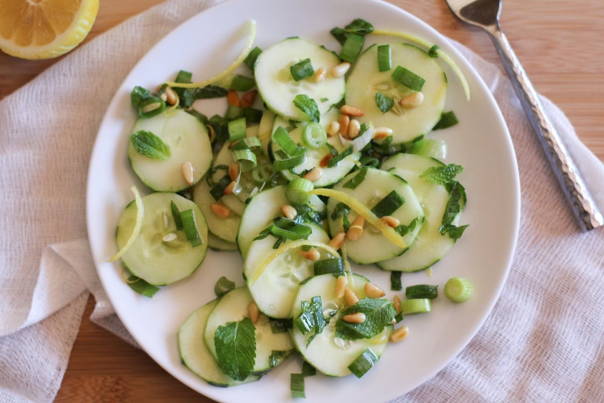 Cucumber salad on a white plate with fresh mint, pine nuts, and more.