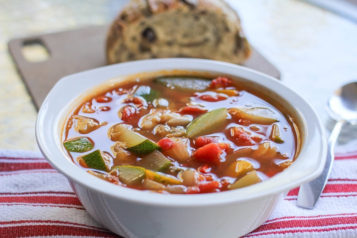 White bowl of zucchini and yellow squash soup with beans and a napkin and spoon to the side.