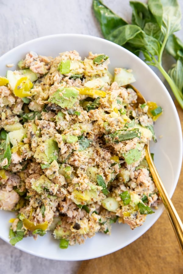 Salmon salad with pickles, avocado, fresh herbs and more mixed up in a white serving bowl with a golden spoon, ready to eat.
