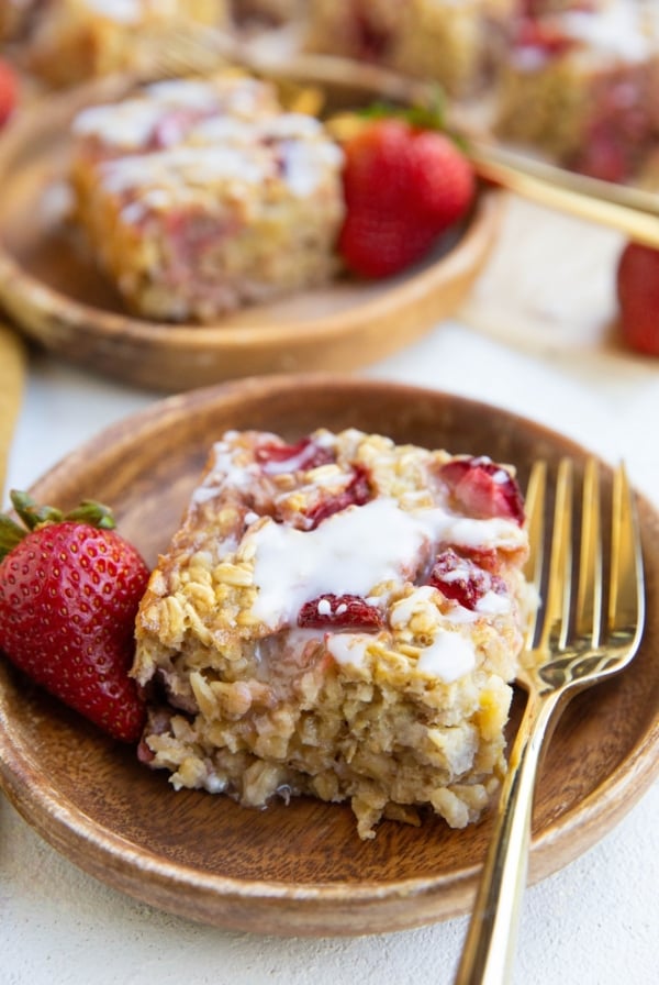 Two wooden plates of strawberry baked oatmeal with a glaze on top and golden forks with fresh strawberries to the side.