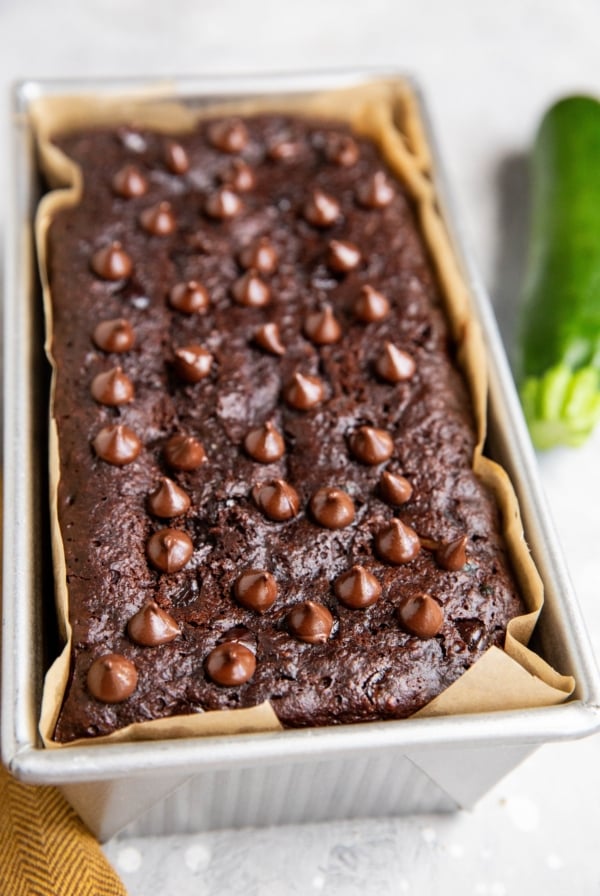 Chocolate zucchini bread in a loaf pan fresh out of the oven with a zucchini to the side.