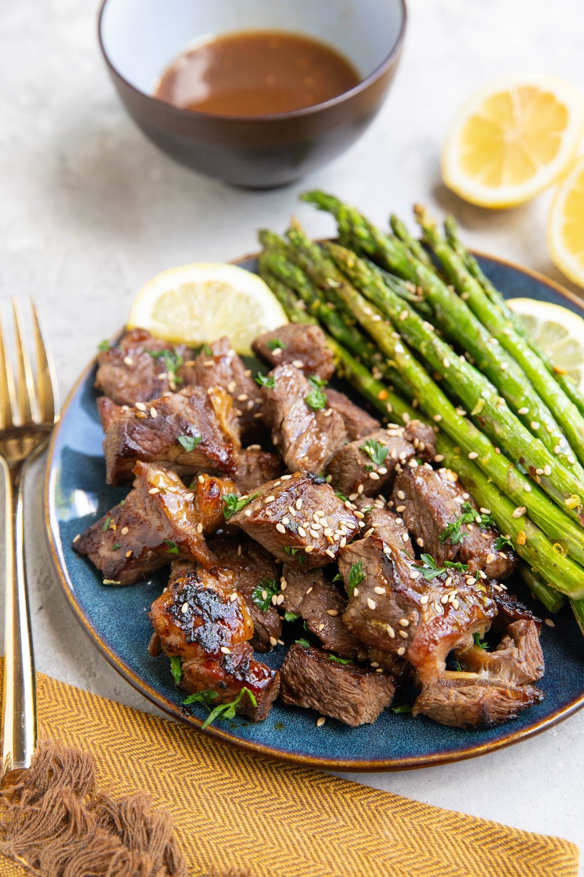 Blue plate with steak bites and asparagus and teriyaki sauce in the background with fresh lemons and a napkin and gold fork.