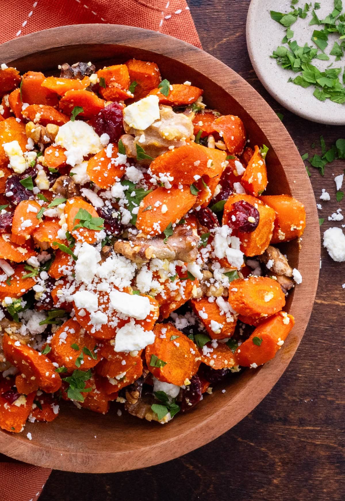 Maple Roasted Carrots in a wooden bowl with feta, dried cranberries and walnuts and a small bowl of parsley to the side.