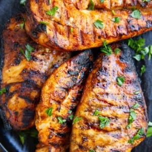 Black plate with four grilled chicken breasts sprinkled with parsley and slices of lemon to the side.