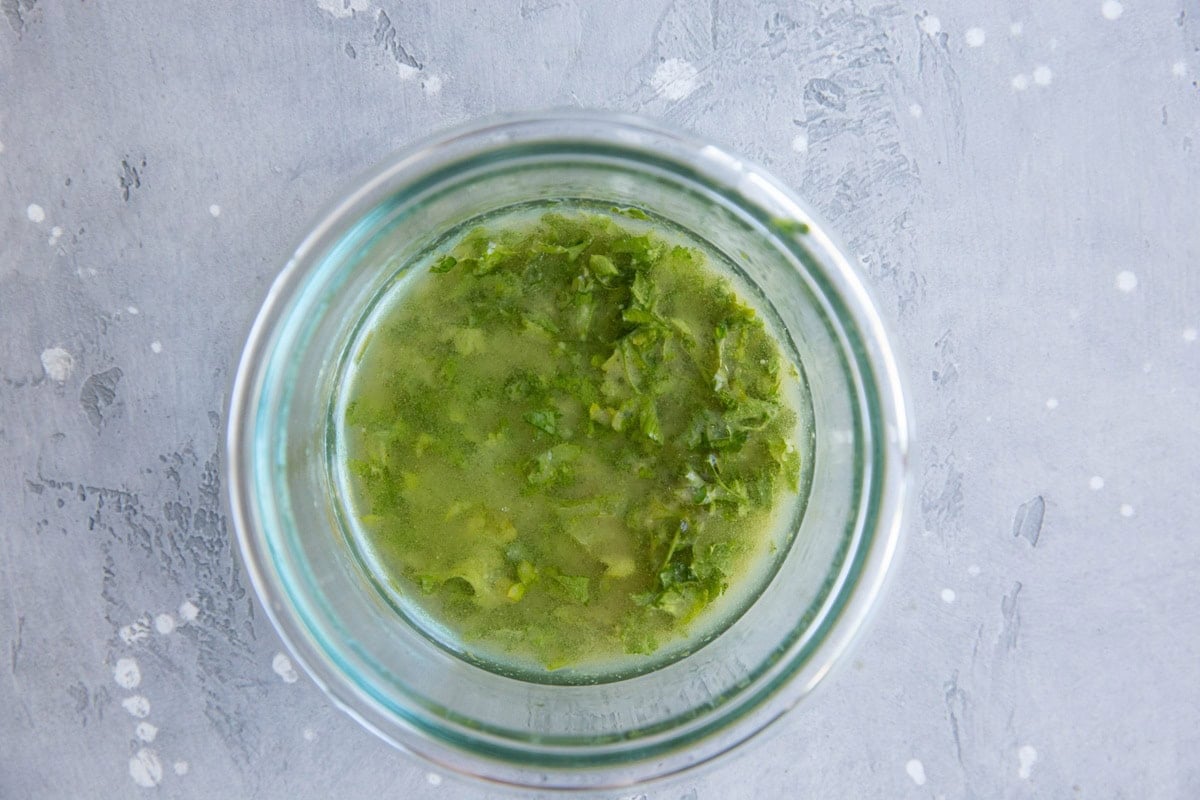 A glass container filled with a green, chunky liquid—perfect as a dressing for a fall harvest salad—sits on a light gray textured surface with white speckles.