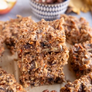Stack of carrot cake breakfast bars on a sheet of parchment paper with the pecans in the background.