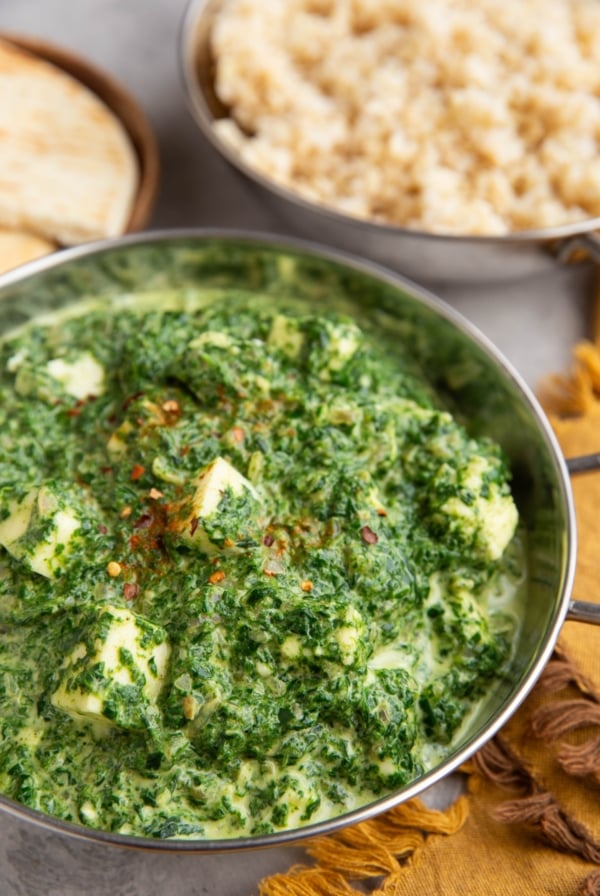 Indian dish full of saag paneer with a dish of brown rice in the background.
