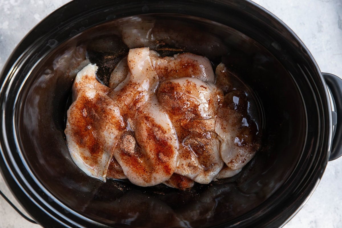Slow cooker full of chicken breasts with spices and oil.