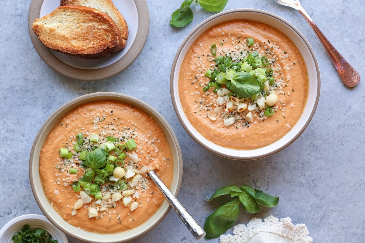 Two bowls of creamy tomato soup with chopped green onions, nuts and seeds sprinkled on top.