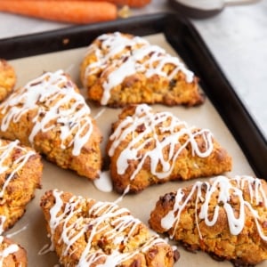 Baking sheet with carrot scones drizzled with glaze with fresh carrots and a mug of coffee in the background.
