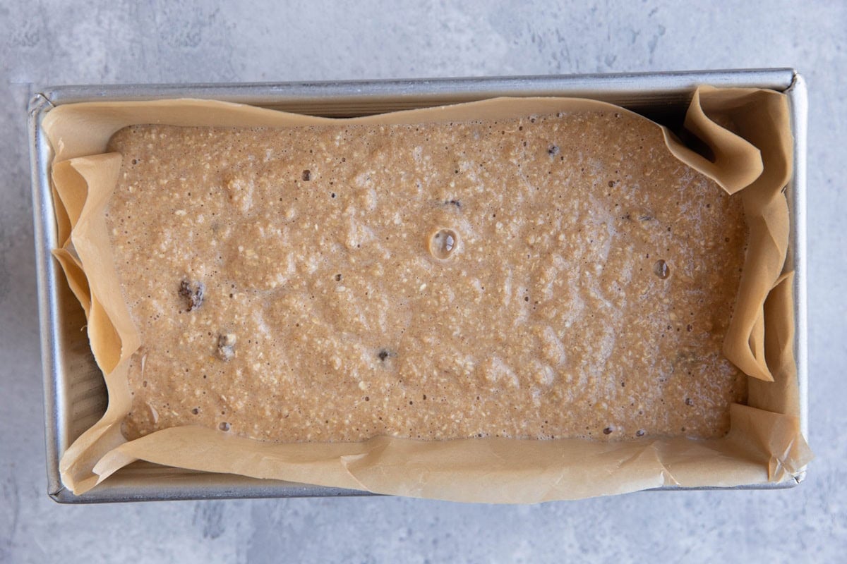 Loaf pan lined with parchment paper with oatmeal raisin bread batter inside.