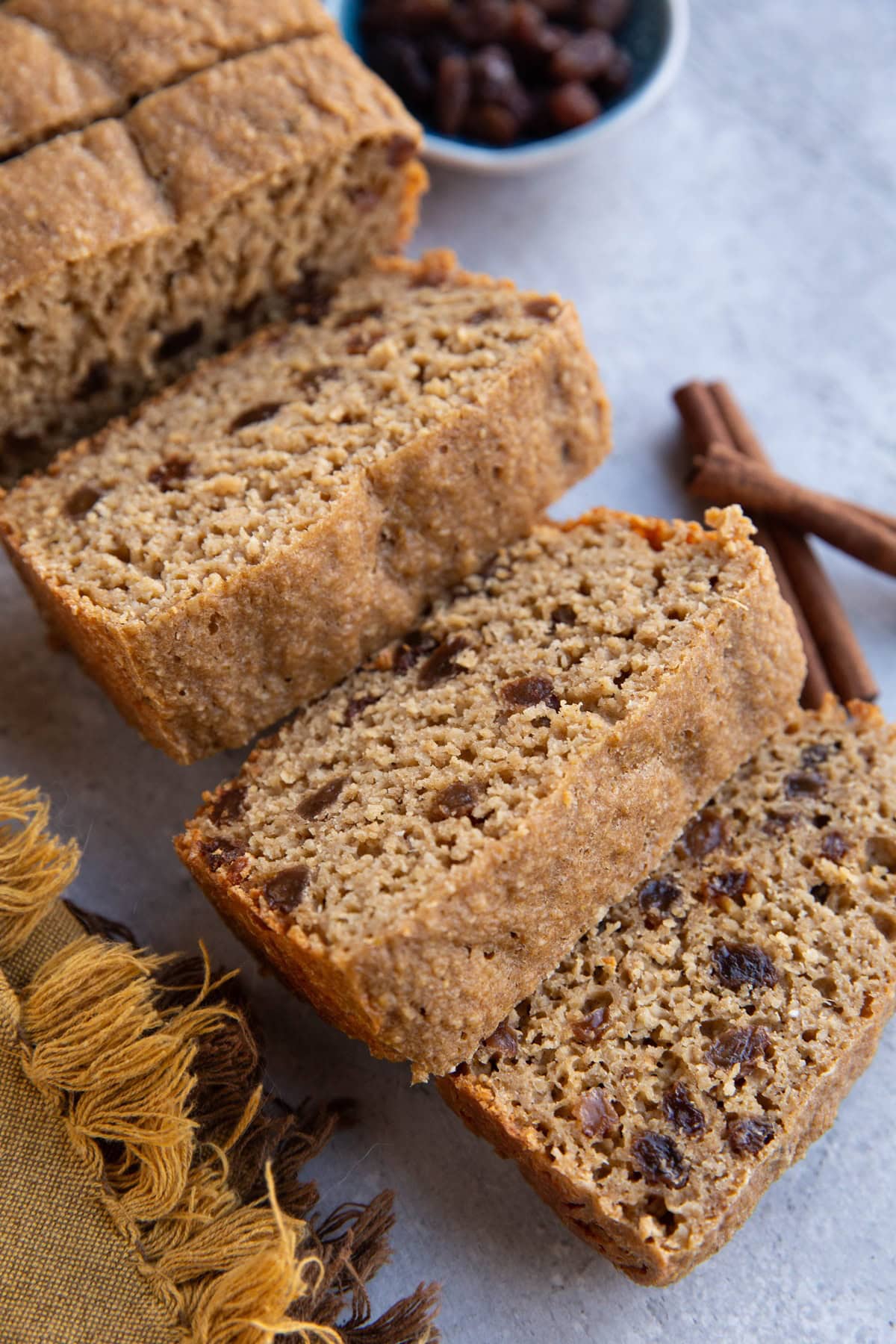 Thick slices of cinnamon raisin bread on a backdrop with raisins and cinnamon sticks in the background.