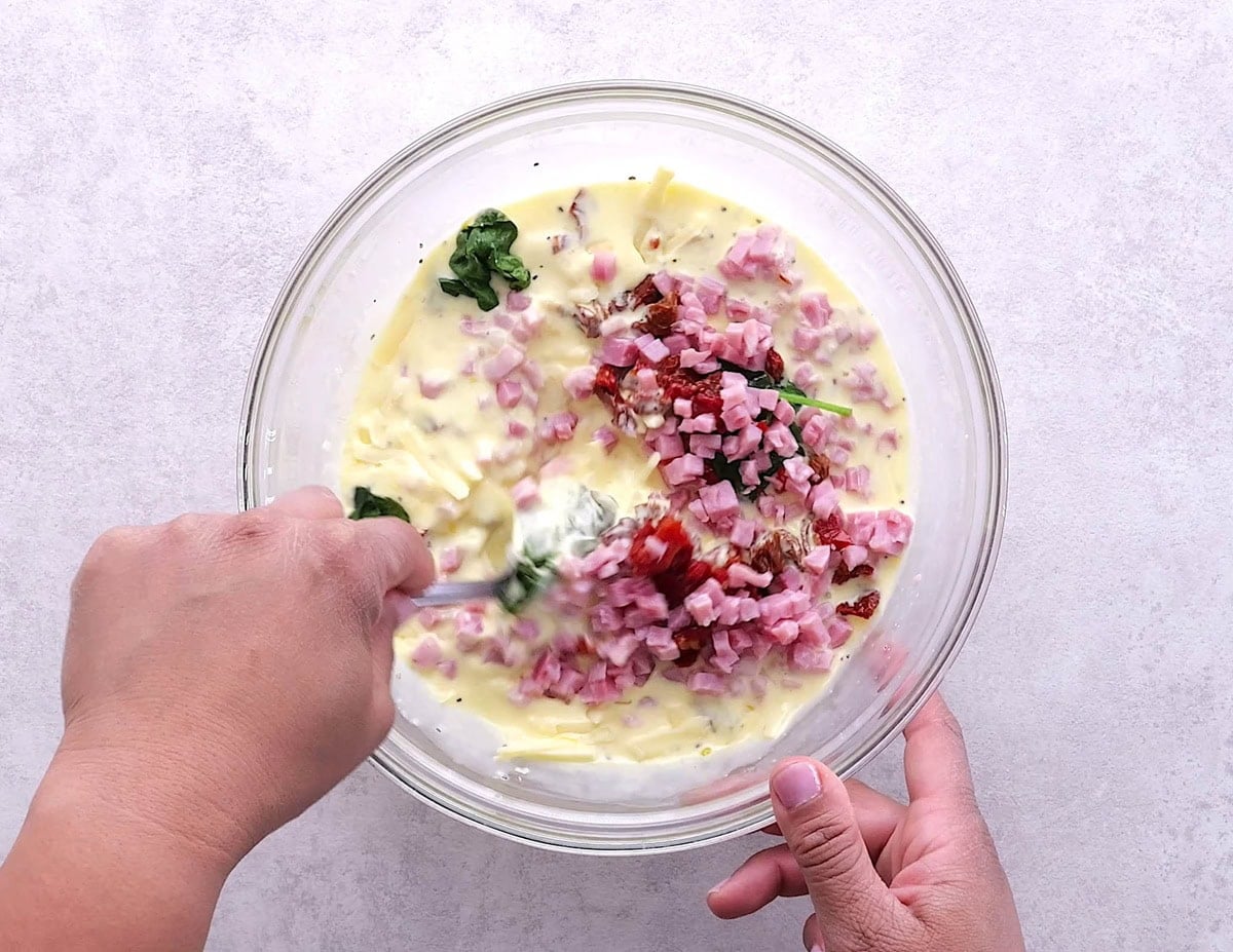 Mixing bowl full of egg custard with spinach, ham, and sun-dried tomatoes being mixed in.