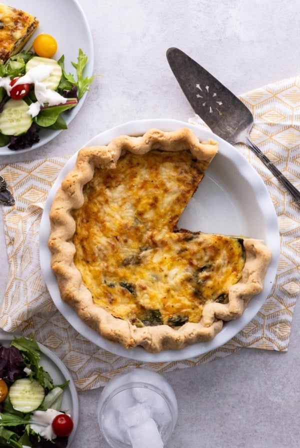 Quiche on a table with a slice taken out and a plate of salad.