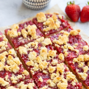 Strawberry crumb bars cut into slices on a sheet of parchment paper with a bowl of strawberries in the background.