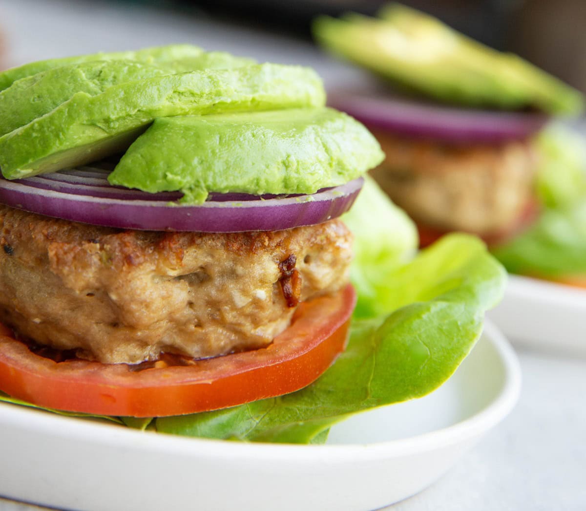 A lettuce-wrapped burger topped with a tomato slice, ground chicken patty, red onion, and sliced avocado, served on a white plate.