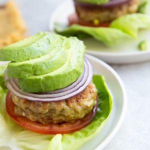 A lettuce-wrapped chicken burger topped with sliced tomato, red onion, and avocado with another chicken patty with toppings in the background.