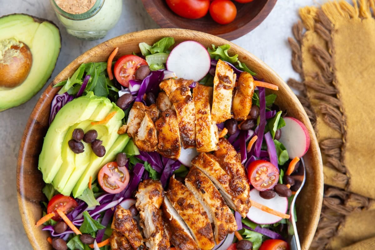 Chicken salad in a wooden bowl with beans, avocado, tomatoes, cabbage, and more. Half of an avocado to the side and a golden napkin.