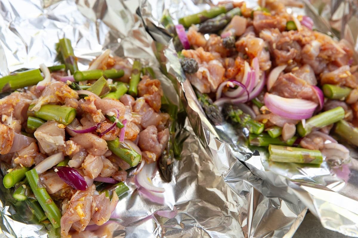 Two long sheets of aluminum foil with chicken, chopped asparagus, and onion tossed in oil and seasonings to make dinner.