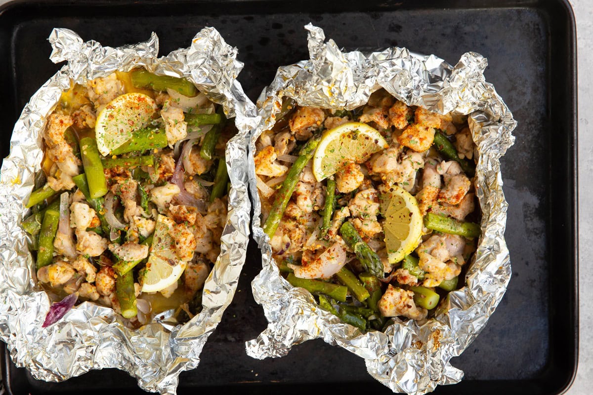 Baking sheet with two packets of aluminum foil with chicken and asparagus.