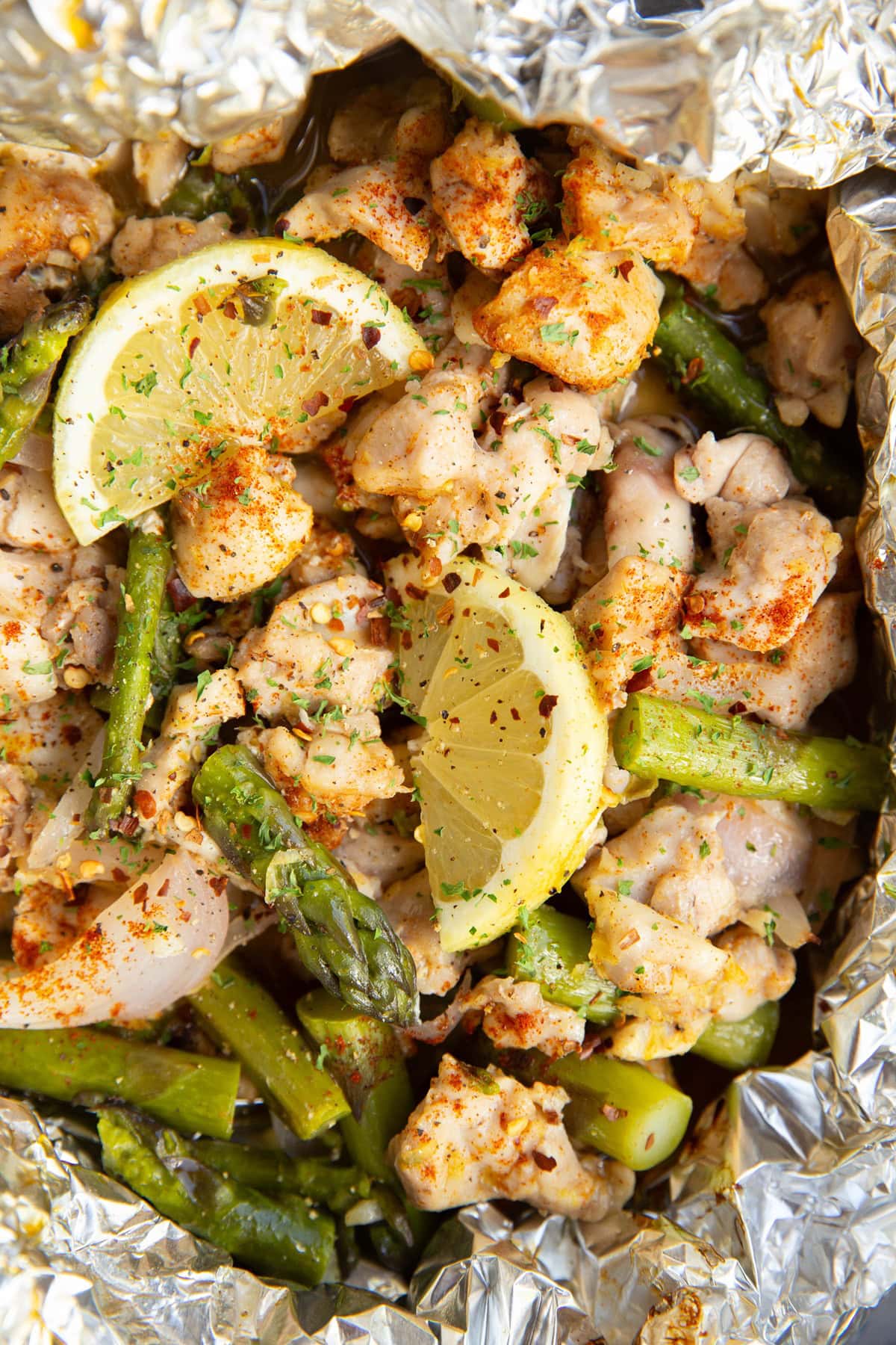 Large packet of foil with cooked chicken, asparagus, and lemon inside, sprinkled with seasonings.