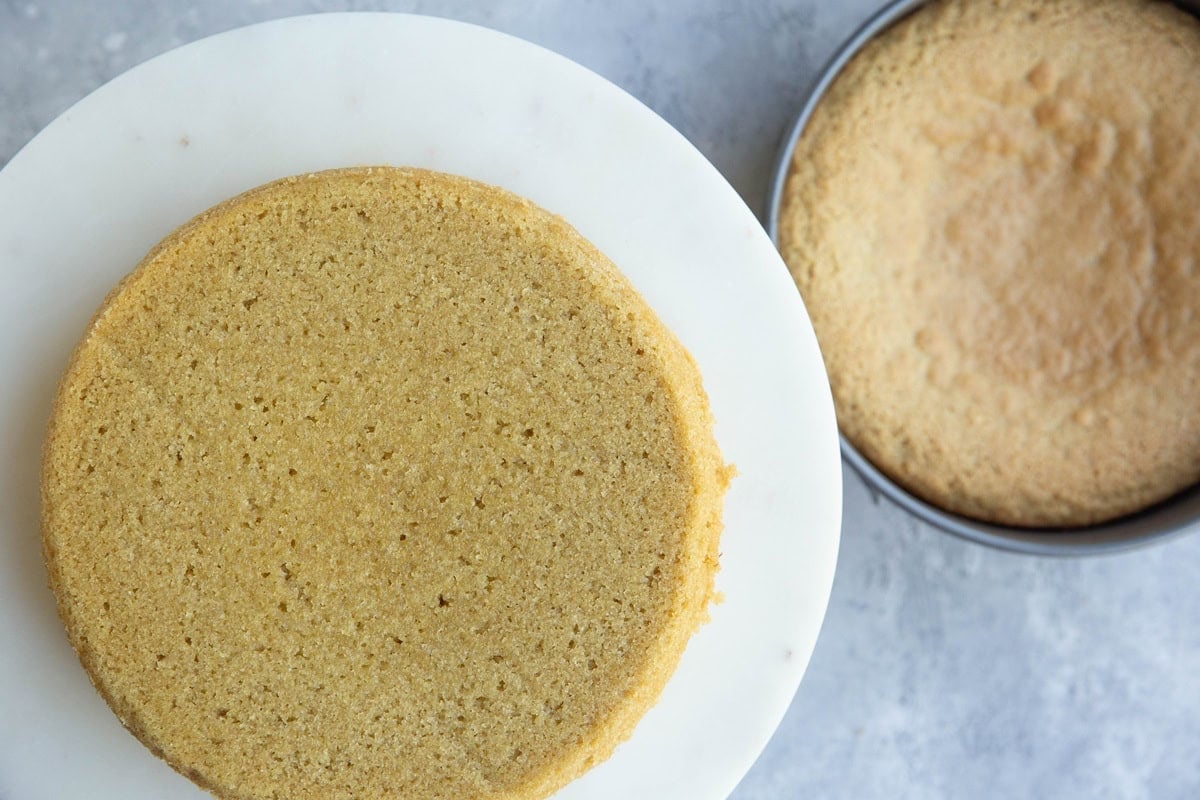 Vanilla cake on a cake stand with another cake still in its pan to the side. Showing how to make a layer cake.