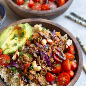 Two large wooden bowls filled with ground turkey mixed with vegetables and sauce on top of cooked brown rice with fresh tomatoes, avocado and cashews. Chop sticks to the side, ready to eat.