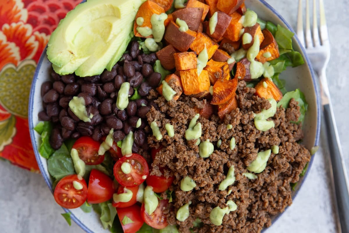 Large bowl full of ground beef, salad greens, rice, tomatoes, avocado, sweet potatoes, and drizzled with creamy avocado dressing.