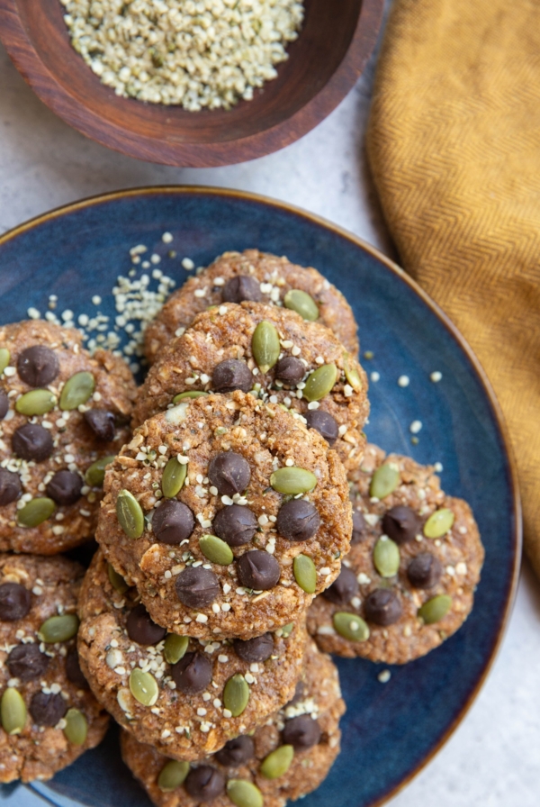 Blue plate of breakfast cookies topped with chocolate chips, pumpkin seeds, and hemp seeds. A bowl of hemp seeds and a golden napkin to the side.