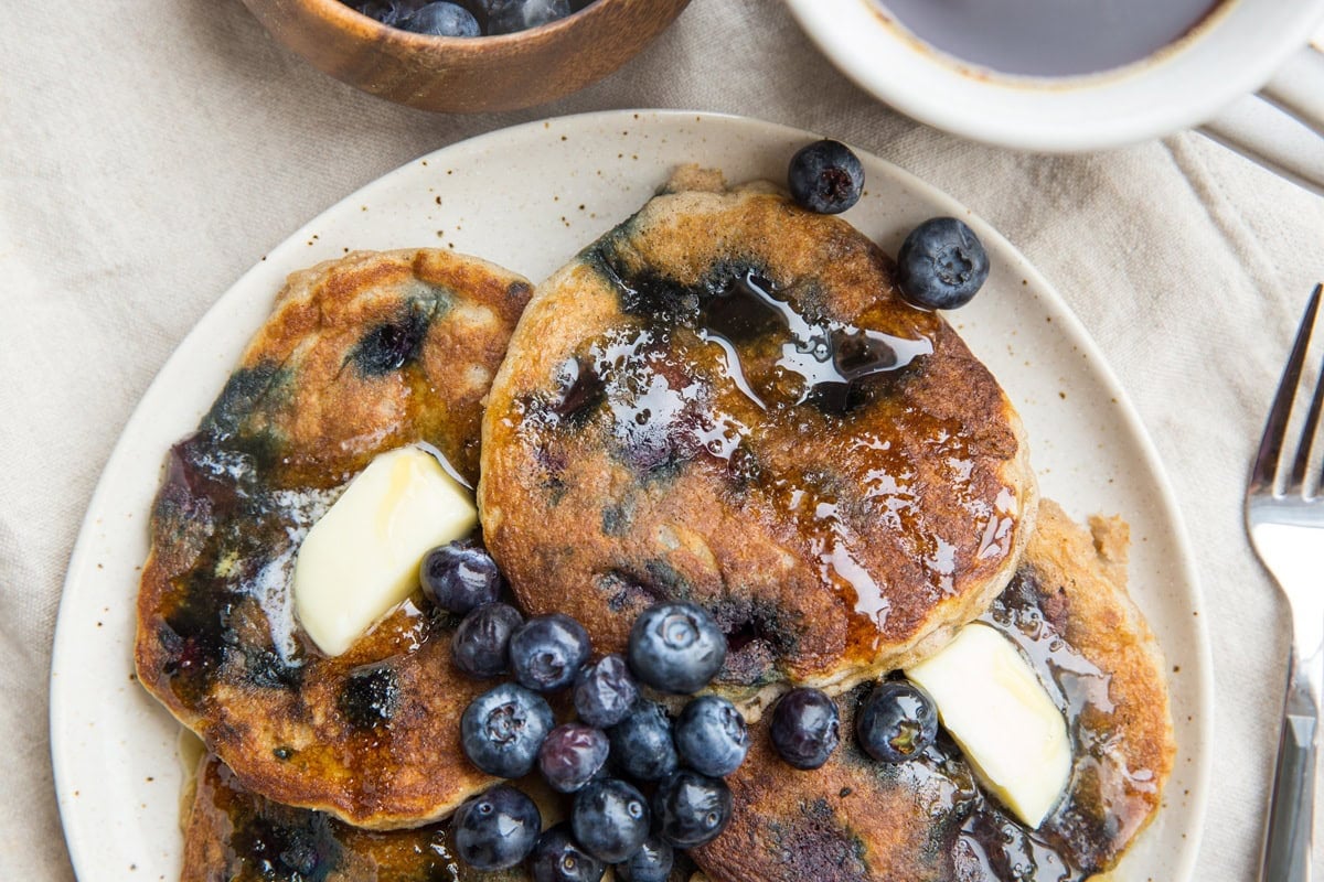 Plate full of almond flour blueberry pancakes drizzled in maple syrup with fresh berries on top and melted butter. A fork to the side, ready to eat.