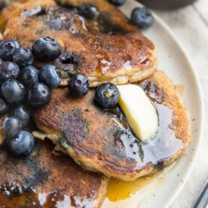 Almond flour pancakes on a plate with fresh blueberries, melted butter, and honey. A fork to the side, ready to serve.