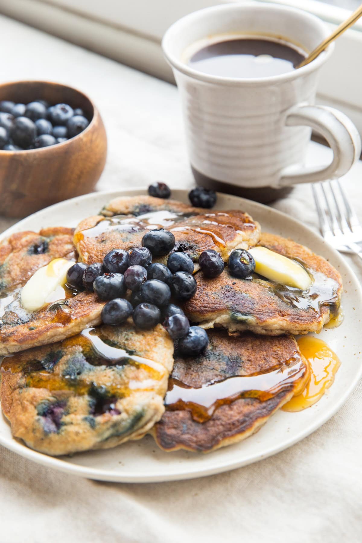 Plate of five blueberry pancakes with melted butter, honey drizzled on top and fresh berries. A mug of coffee and a bowl of blueberries in the background.
