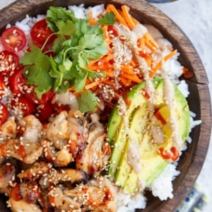 Big wooden bowl full of grilled chicken, rice, avocado, carrots, cherry tomatoes, drizzled with peanut sauce and sriracha. A bowl of peanut sauce and chopsticks to the side.