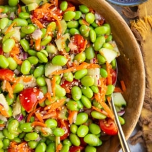 Edamame salad in a wooden bowl with a golden spoon and a blue bowl of sesame seeds to the side.