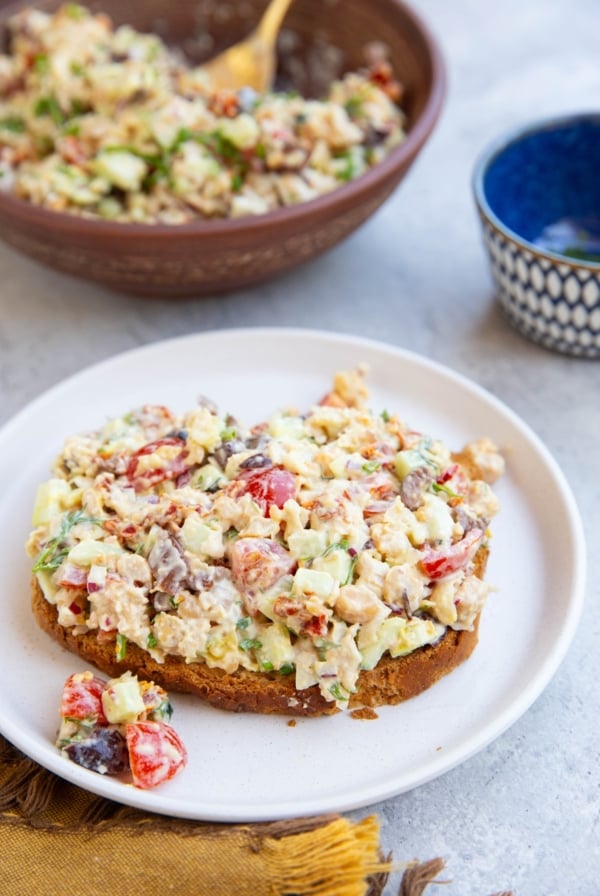 Bread with chickpea tuna salad on top on a white plate and the rest of the salad in a bowl in the background. A golden napkin to the side.