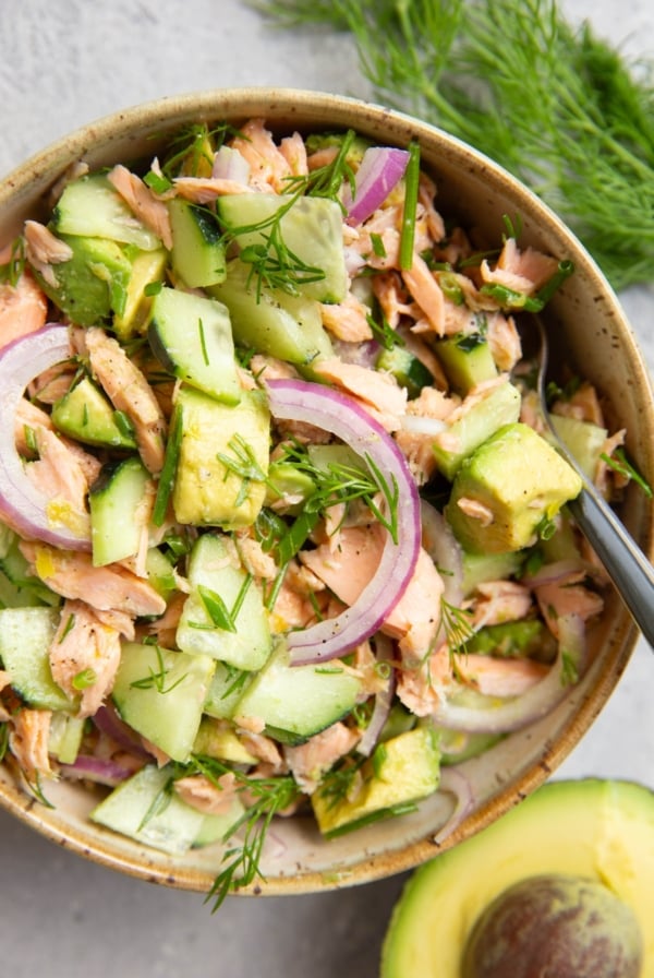 Bowl of cucumber dill avocado salmon salad with a fork ready to serve, half an avocado to the side and fresh dill to the side.