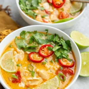 Two bowls of coconut lime fish soup with lime wedges, cilantro, and red chili peppers on top with a cut lime to the side and a golden napkin.