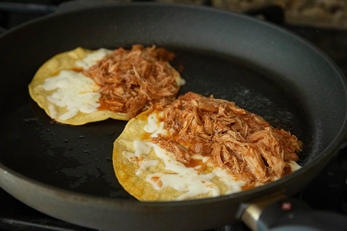 Two tacos with corn tortillas, cheese, and chicken cooking on a skillet to become crispy.