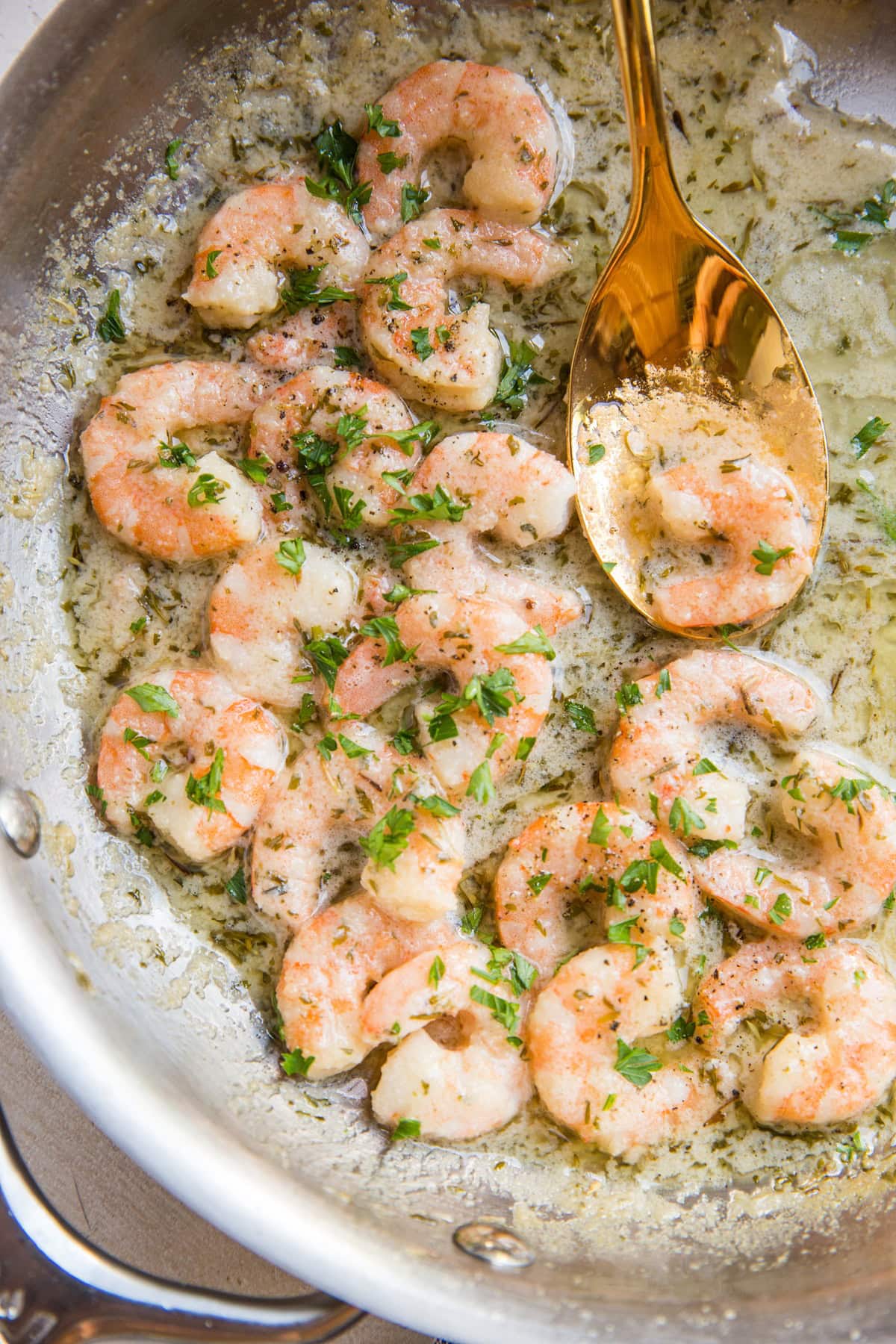 Stainless steel skillet full of sauteed shrimp in a lemon butter garlic sauce with a golden spoon scooping up one of the shrimp.