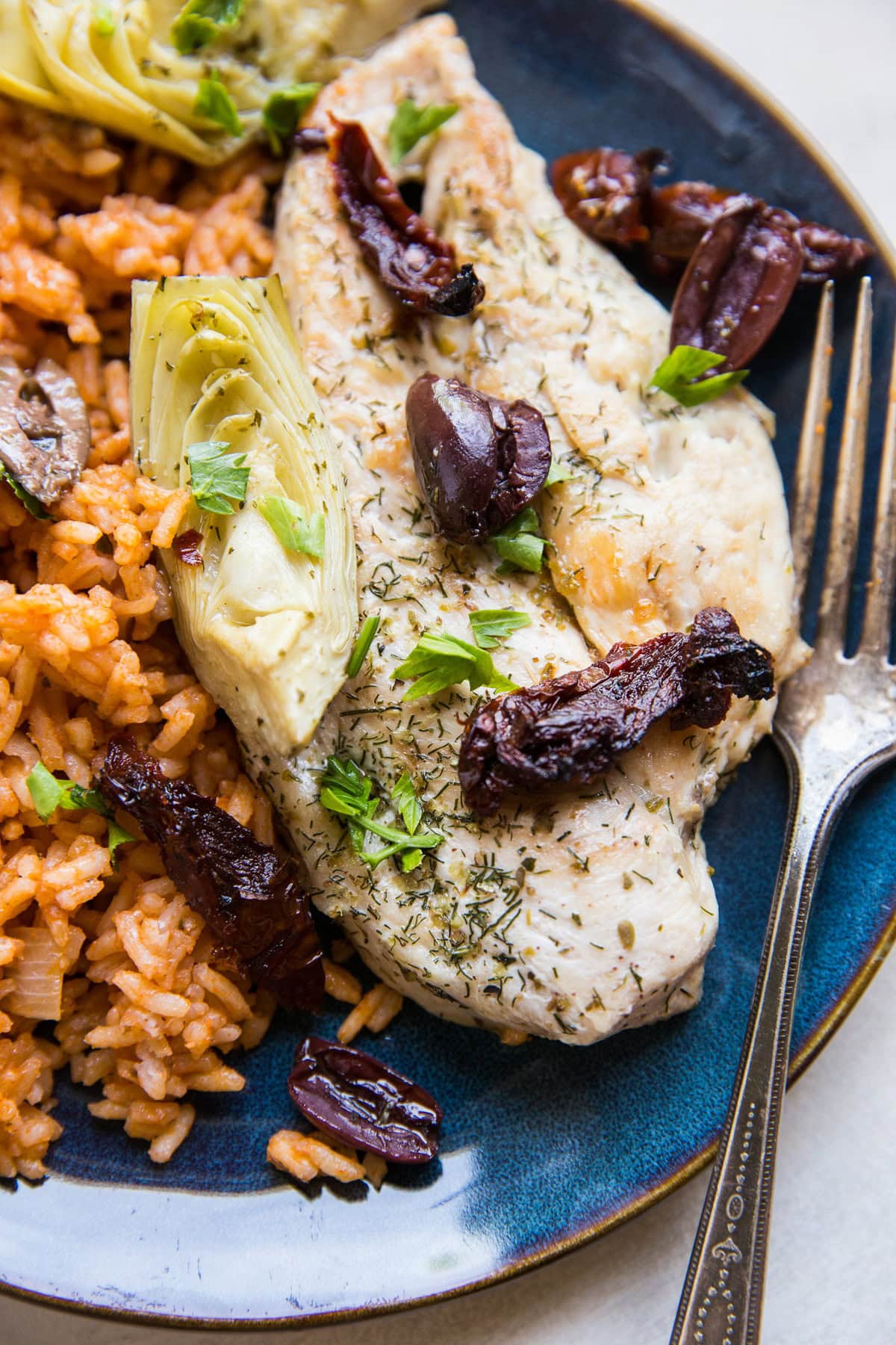 Chicken breast on a plate with sun-dried tomatoes, kalamata olives, and artichoke hearts on top. Next to the chicken is Spanish rice.
